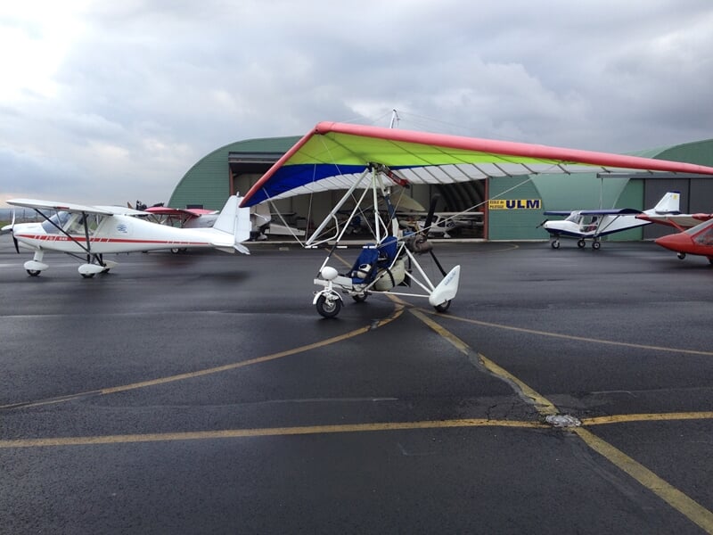 Plusieurs ULM stationnés sur une piste humide devant des hangars verts sous un ciel nuageux.