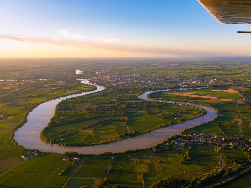 Vue aérienne au crépuscule des méandres de la dordogne traversant des paysages verdoyants, avec une ville en arrière-plan et l'aile d'un avion dans le coin supérieur droit