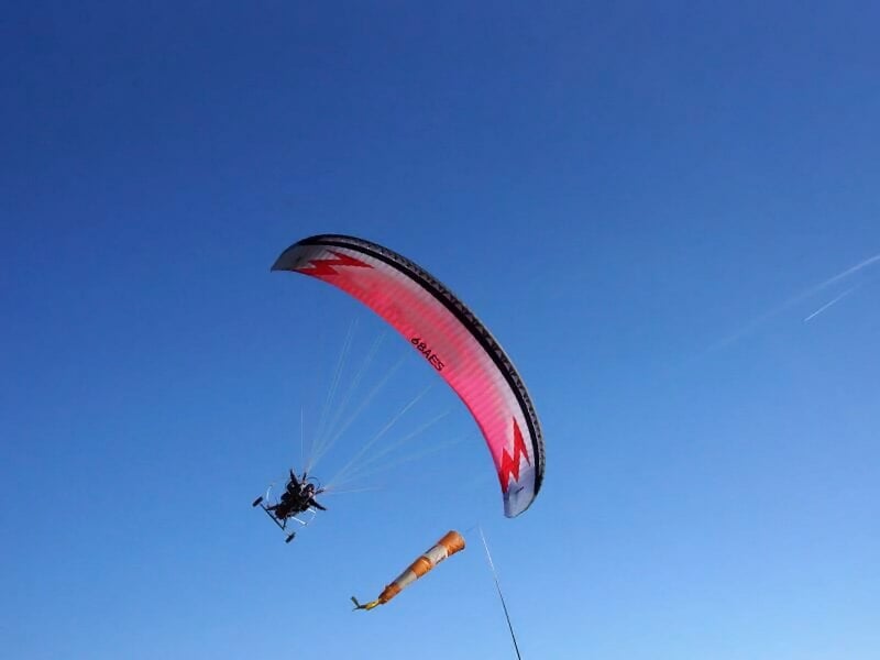 Paramoteur rouge volant dans un ciel bleu clair au-dessus d'un paysage naturel avec un anémomètre orange.