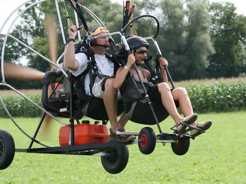 Deux personnes en tandem sur un paramoteur au sol, prêtes à décoller sur un champ vert.