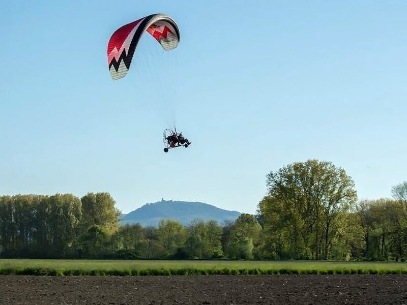 Paramoteur rouge volant au-dessus d'un champ avec des arbres et une colline en arrière-plan.