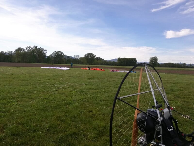 Paramoteur au sol sur un champ vert avec des parapentes déployés en arrière-plan.