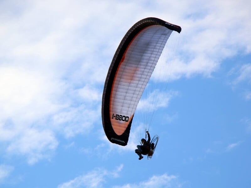 Paramoteur en vol avec une voile blanche et noire contre un ciel bleu clair avec quelques nuages.