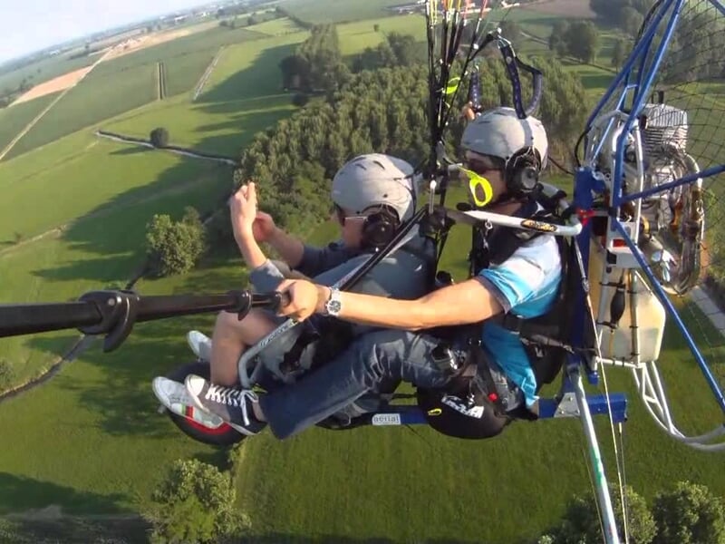 Deux pilotes en paramoteur volant au-dessus d'une campagne verte avec des arbres et des champs en arrière-plan.