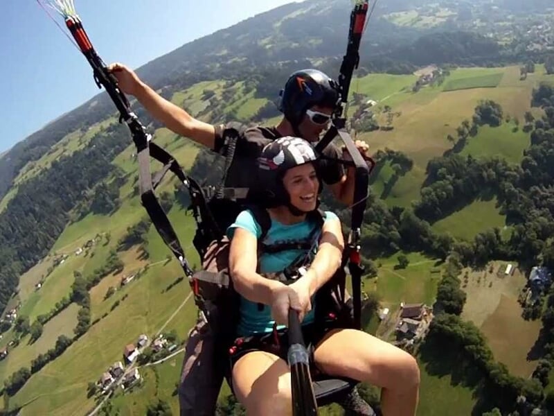 Femme souriante en tandem paramoteur avec un instructeur, survolant une zone verte et vallonnée.