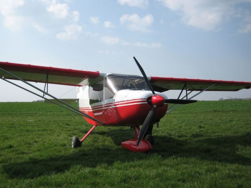 Avion léger rouge et blanc à hélice avant, stationné sur un champ d'herbe sous un ciel partiellement nuageux