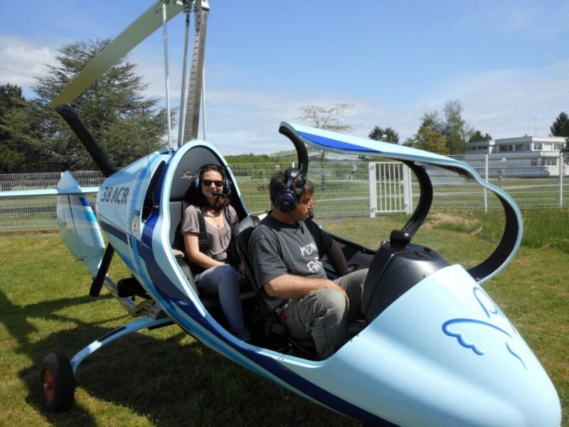 Autogire bleu avec numéro 38 ACR, deux passagers portant des casques audio, prêts pour un vol, stationné sur une pelouse ensoleillée près de bâtiments modernes