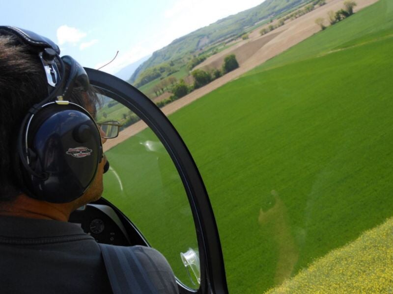 Vue aérienne d'un champ verdoyant depuis l'intérieur d'un ULM autogire, avec un pilote portant un casque audio, survolant une vallée par temps ensoleillé