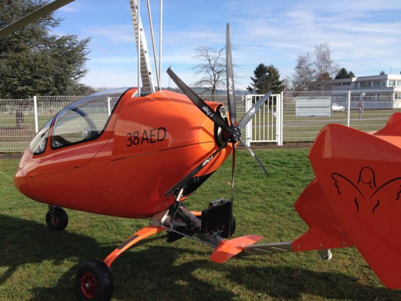 ULM autogire orange avec numéro 38 AED, hélice arrière et cockpit vitré, stationné sur une pelouse, près d'une clôture blanche et de bâtiments