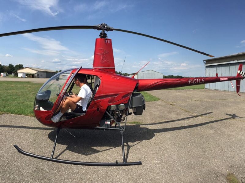 Hélicoptère rouge au sol avec un pilote assis dans le cockpit prêt à décoller sur une piste d'aviation.