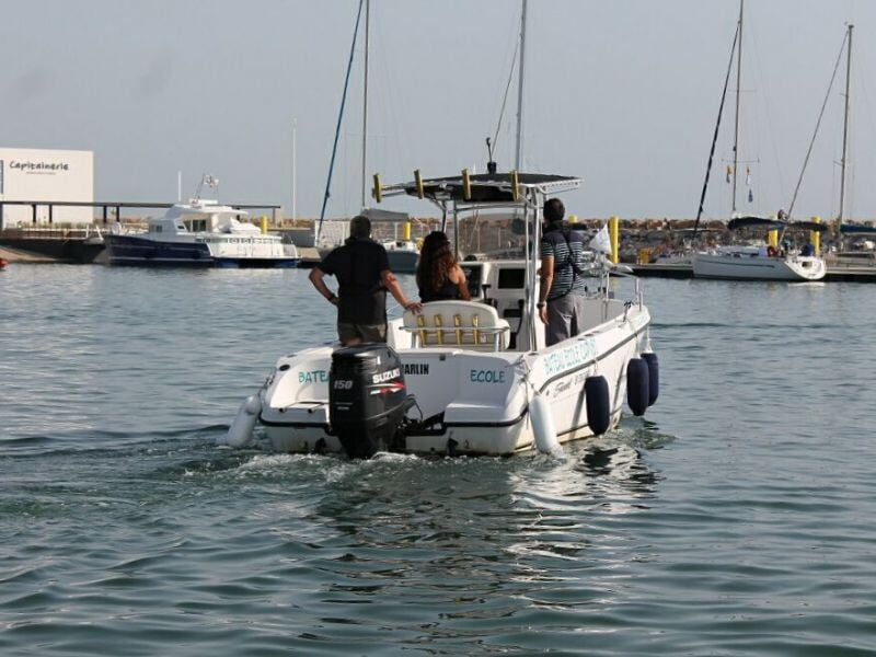 Un bateau blanc avec trois personnes à bord navigue dans un port calme avec plusieurs autres bateaux amarrés.