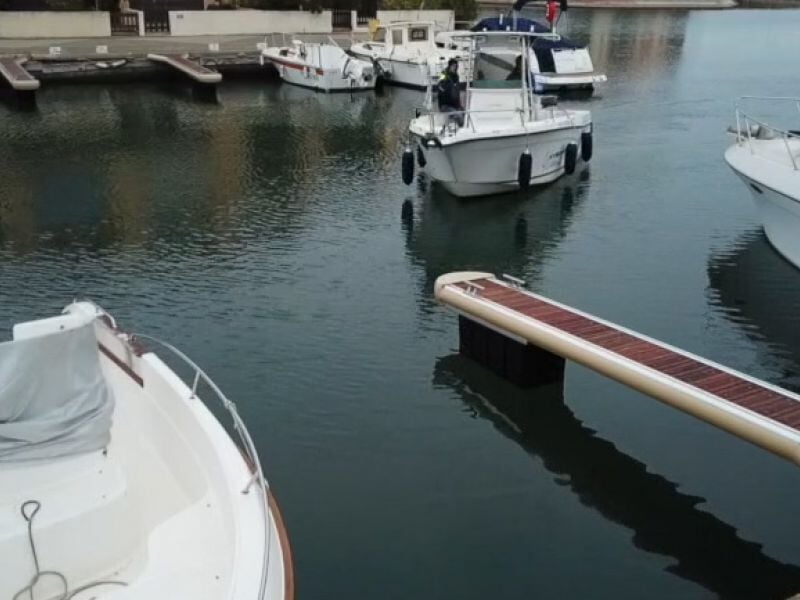 Vue d'un petit port avec plusieurs bateaux blancs amarrés, un ponton en bois s'avance sur l'eau calme.