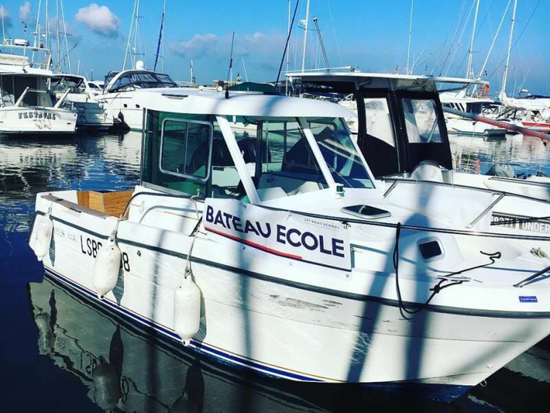 Bateau école blanc amarré dans un port avec plusieurs autres bateaux et un ciel bleu clair en arrière-plan.