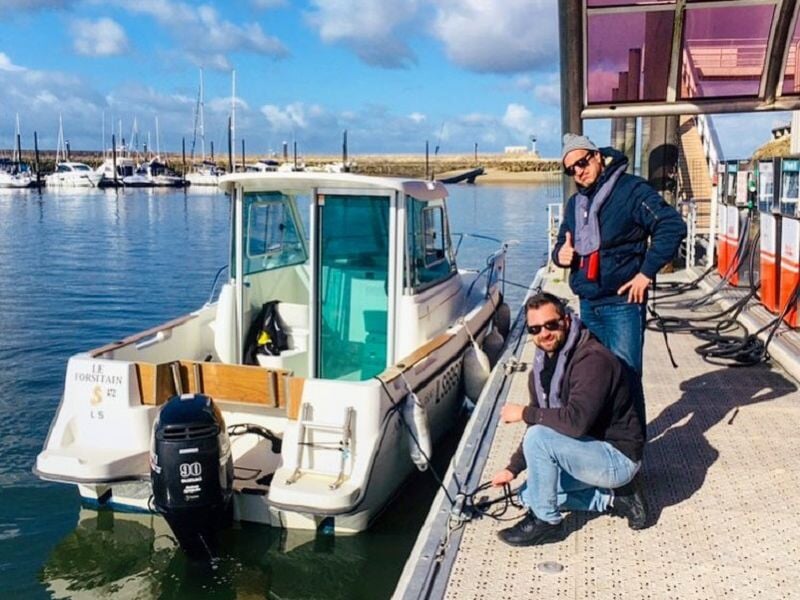 Deux hommes posent à côté d'un bateau blanc amarré dans un port avec des bâtiments et des bateaux en arrière-plan.