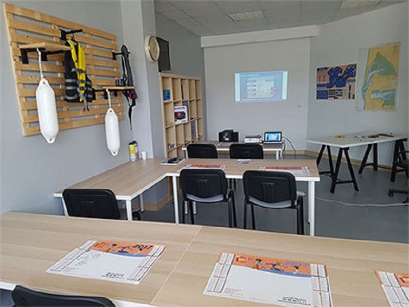 Salle de classe avec tables, chaises, matériel nautique accroché au mur et un écran de projection allumé.