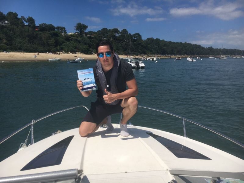 Homme accroupi sur un bateau blanc tenant un livret, avec des bateaux et une plage en arrière-plan.