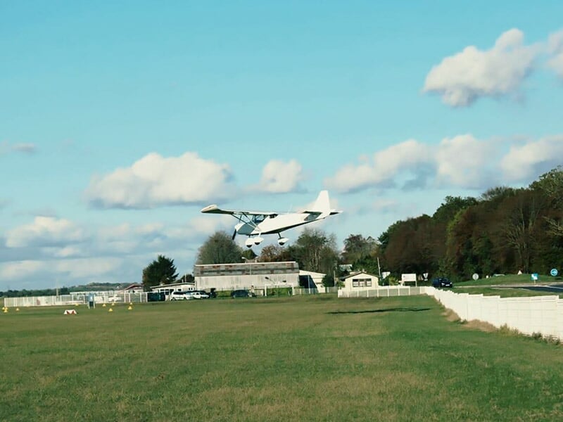 Petit avion blanc en train de décoller ou d'atterrir sur une piste herbeuse avec un ciel bleu et nuageux.