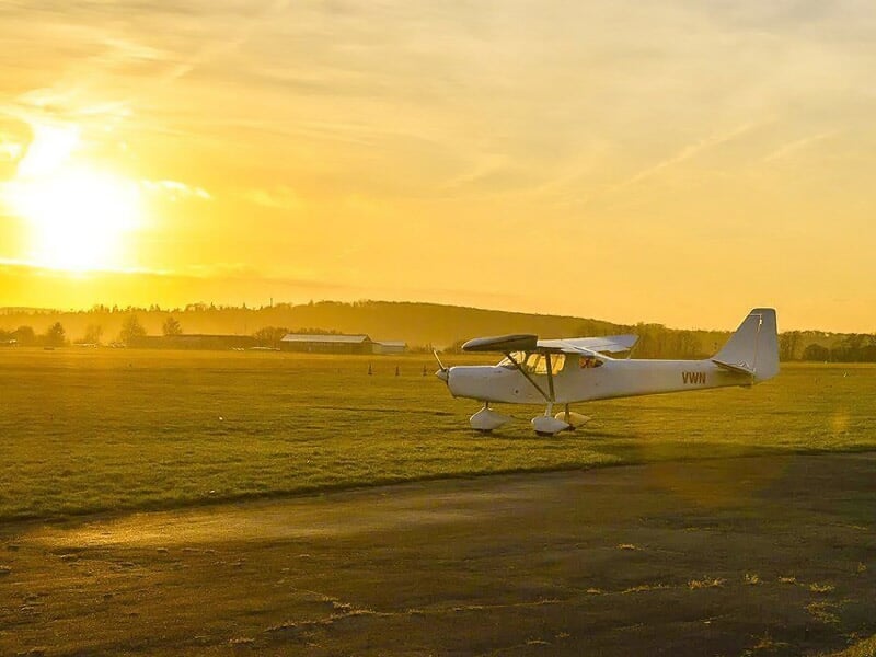 Avion léger blanc sur une piste d'herbe au coucher du soleil avec un ciel orange et paysage vallonné.