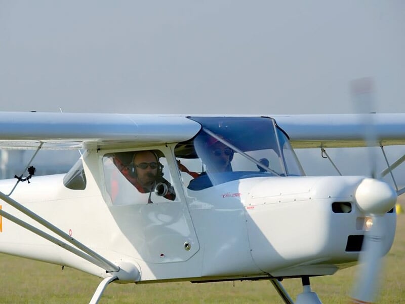 Pilote dans un petit avion blanc avec hélice en mouvement, posé sur une piste d'herbe verte.
