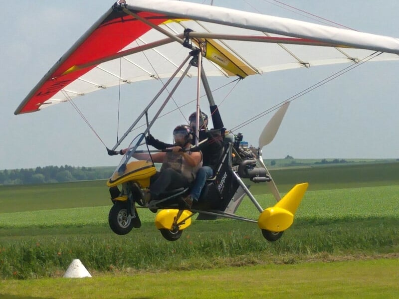 ULM jaune et rouge avec deux personnes à bord volant au-dessus d'un champ vert sous un ciel clair.