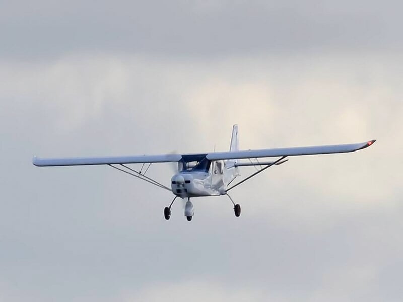 Petit avion blanc volant dans un ciel nuageux avec train d'atterrissage visible en dessous.