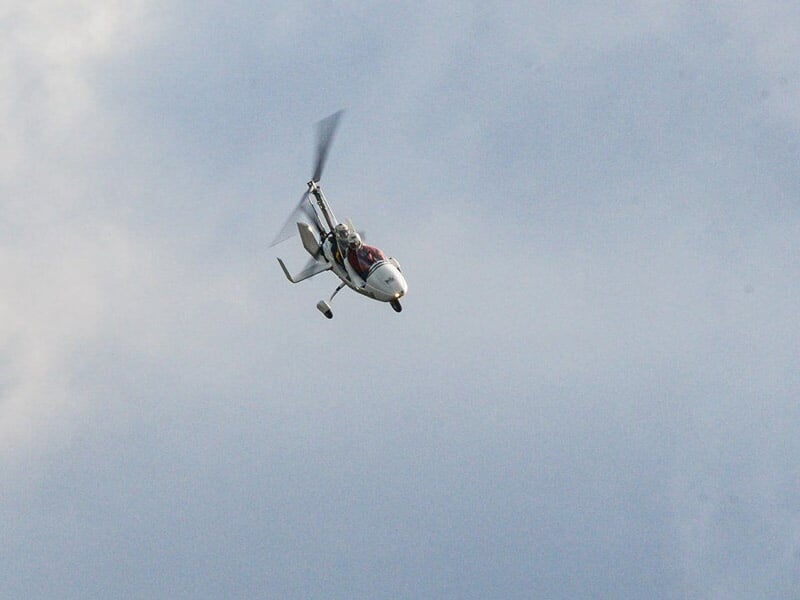 Gyrocoptère blanc avec deux passagers volant dans un ciel gris clair au-dessus d'un paysage flou.