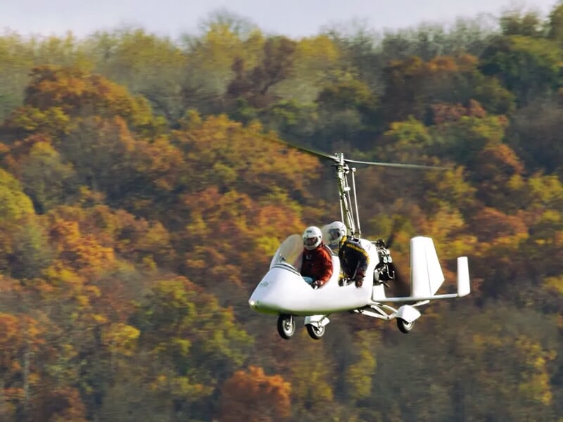 Gyrocoptère blanc avec deux personnes à bord volant devant un fond d'arbres aux couleurs d'automne.