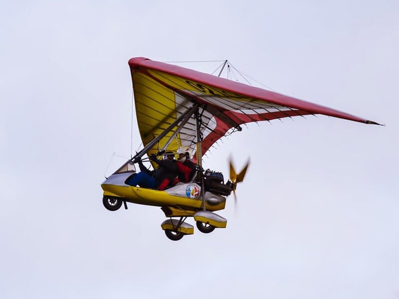 ULM jaune et rouge avec deux personnes à bord volant au-dessus d'un champ vert sous un ciel nuageux.