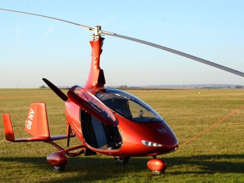 Gyrocoptère rouge stationné sur une piste d'herbe verte avec un ciel bleu clair en arrière-plan.