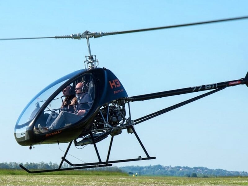 Petit hélicoptère noir avec deux personnes à bord volant au-dessus d'un champ vert sous un ciel clair.