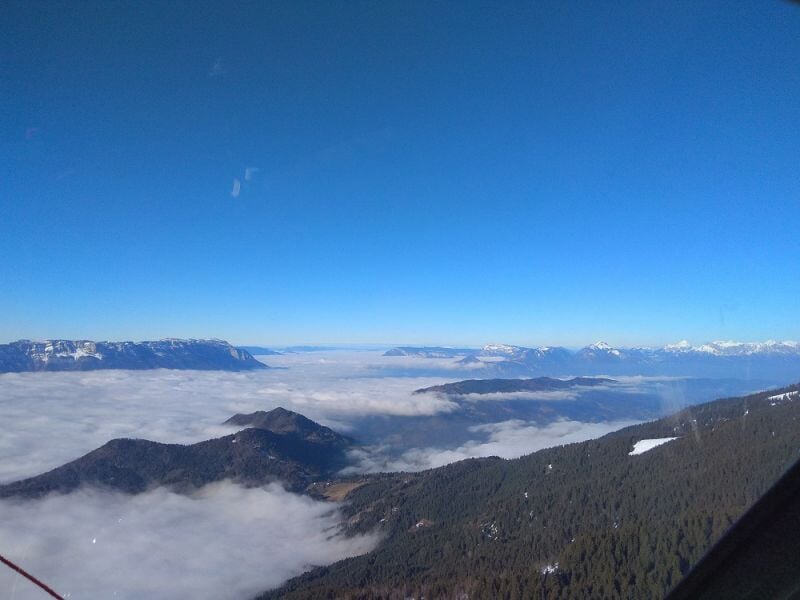 Vue aérienne d'un paysage montagneux avec des nuages bas et un ciel bleu clair.