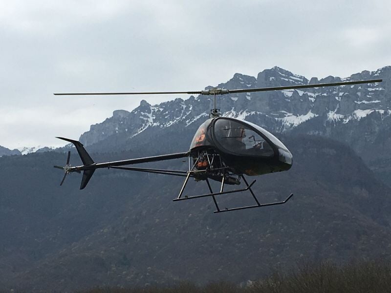 Autogire volant devant une chaîne de montagnes rocheuses sous un ciel nuageux.