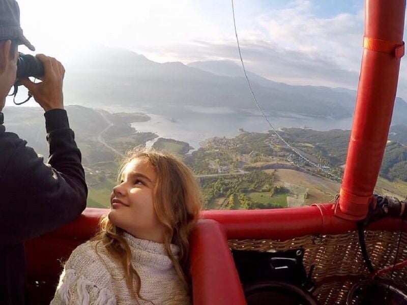 Une jeune fille souriante regarde un homme prenant une photo depuis une montgolfière au-dessus d'un paysage montagneux.