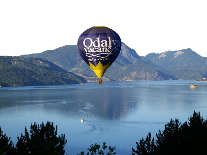 Montgolfière jaune avec inscription Odal Vacances survolant un lac entouré de montagnes sous un ciel clair.