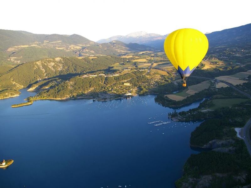 Montgolfière jaune volant au-dessus d'un grand lac entouré de collines et de champs ensoleillés.