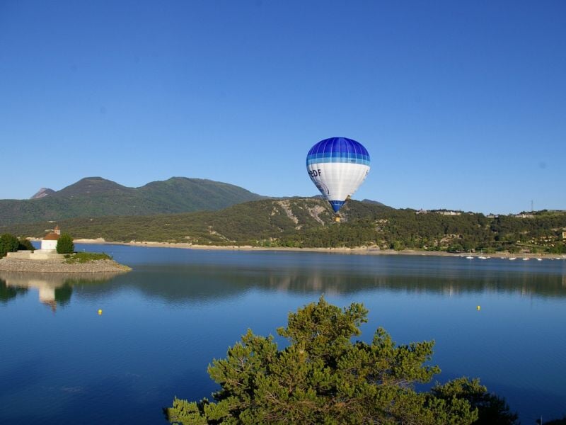 Montgolfière bleue et blanche survolant un lac calme avec des collines et un ciel bleu clair en arrière-plan.