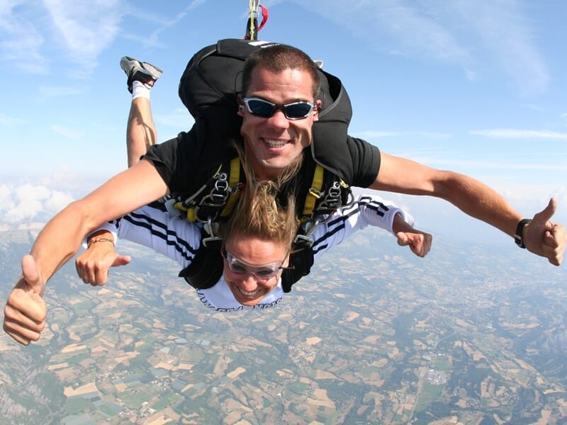 Un saut en tandem en parachute, avec l'instructeur et le passager donnant un pouce en l'air, montrant des émotions positives, au-dessus d'un paysage parsemé de nuages et de terres verdoyantes