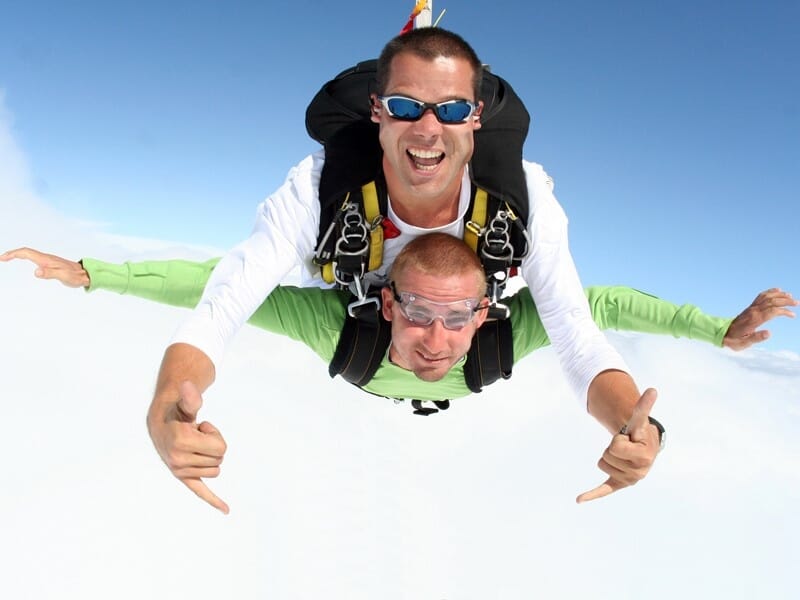 Deux parachutistes en tandem en chute libre, l'instructeur et le passager font des signes devant un fond de nuages blancs éclatants et un ciel bleu clair
