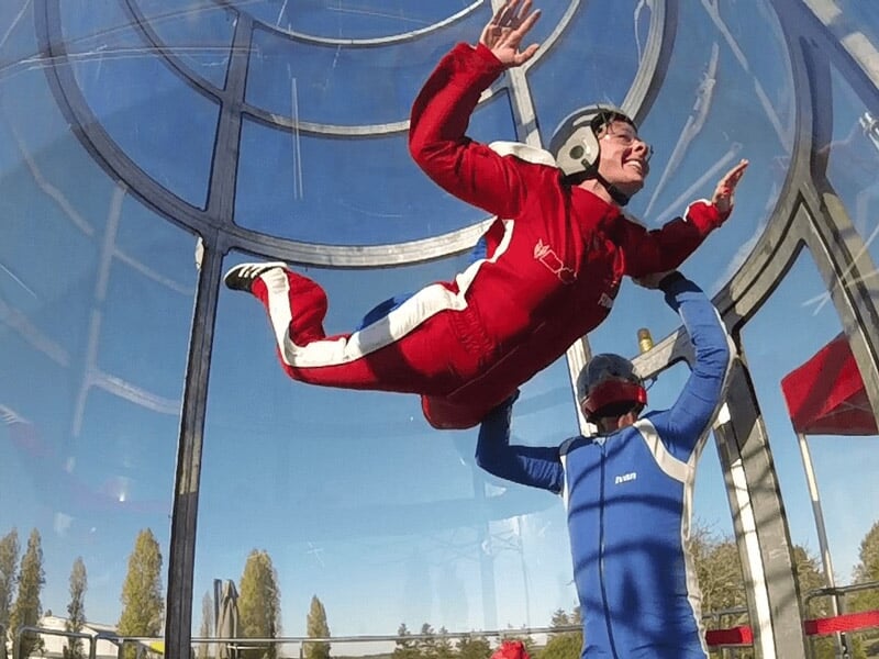 Vol simulé dans un tunnel de vent, participant en combinaison rouge levé par un instructeur en bleu, arrière-plan avec arbres et ciel dégagé.
