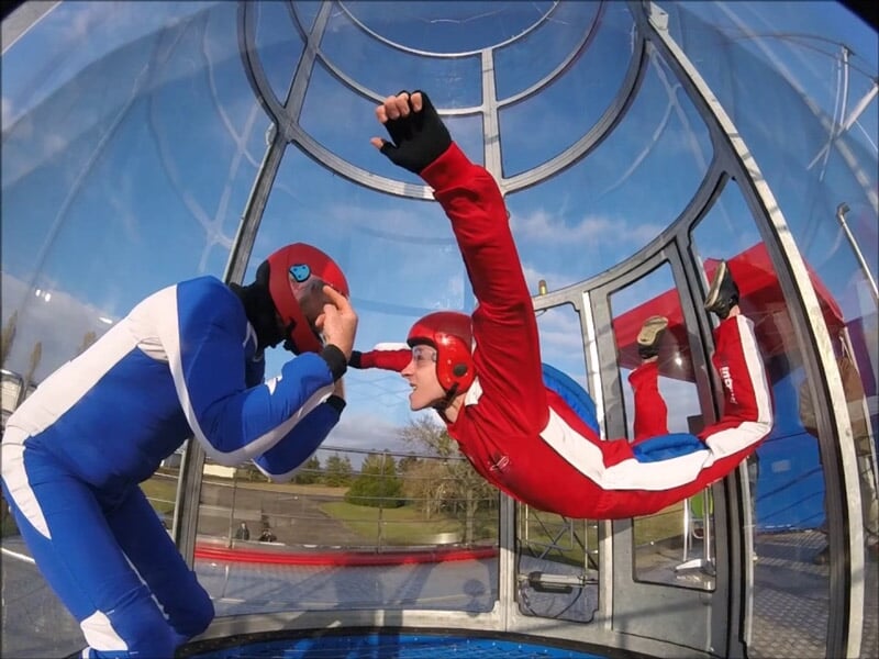 Interaction entre un instructeur en combinaison bleue et un participant en rouge dans un simulateur de chute libre, posture de vol horizontale avec casque rouge et protection oculaire.
