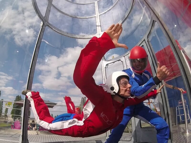 Enfant en vol horizontal dans une soufflerie avec instructeur, tunnel transparent et ciel dégagé.