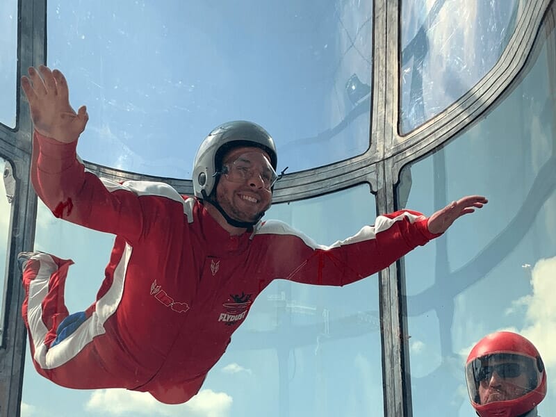 Homme en tenue de vol rouge et blanche lévitant dans un simulateur de chute libre, casque gris, expression concentrée, encadré par des parois vitrées sous un ciel clair.