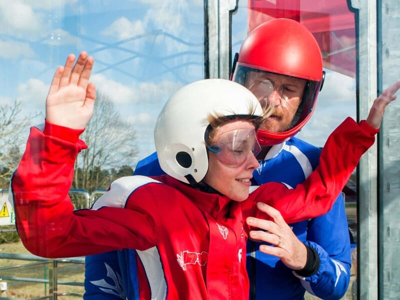 Enfant en combinaison rouge guidé par un instructeur dans une soufflerie vitrée sous un ciel ensoleillé.