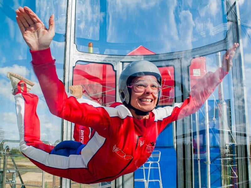 Personne souriante en combinaison rouge et blanche flottant dans un tunnel de vent transparent, casque gris et lunettes de protection, ciel bleu en arrière-plan.