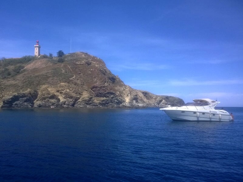 Un yacht blanc au mouillage près d'un littoral rocheux avec un phare blanc et rouge sur une colline en arrière-plan sous un ciel bleu