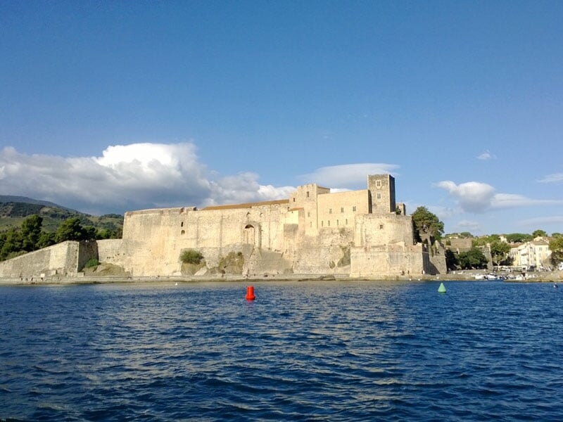 Vue maritime d'une forteresse médiévale massif avec une tour carrée dominant une ville côtière sous un ciel partiellement nuageux