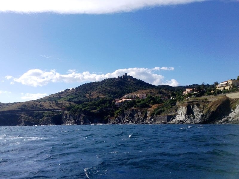 Vue depuis la mer d'une colline avec des bâtiments méditerranéens et une forteresse au sommet, dominant une côte rocheuse sous un ciel bleu avec des nuages