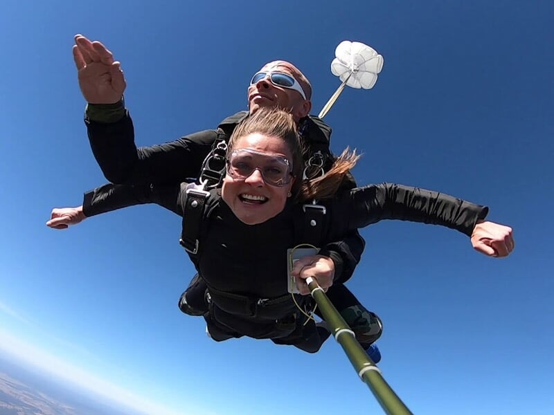 Chute libre en tandem avec une femme souriante et un instructeur, petit parachute de freinage visible, ciel bleu intense en arrière-plan