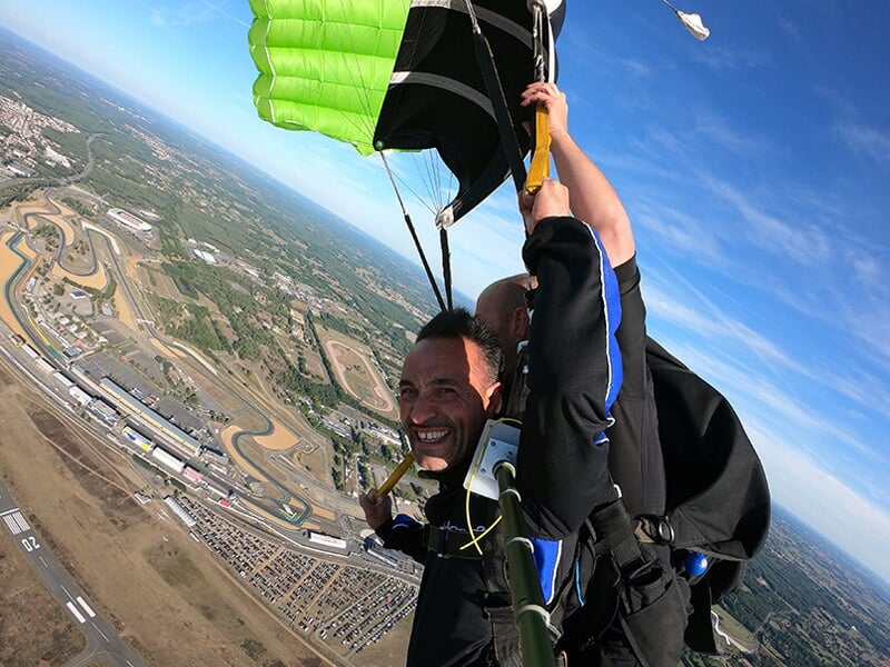 Parachutisme tandem avec un parachute vert déployé, large sourire du participant, panorama d’un circuit automobile et de terrains en arrière-plan