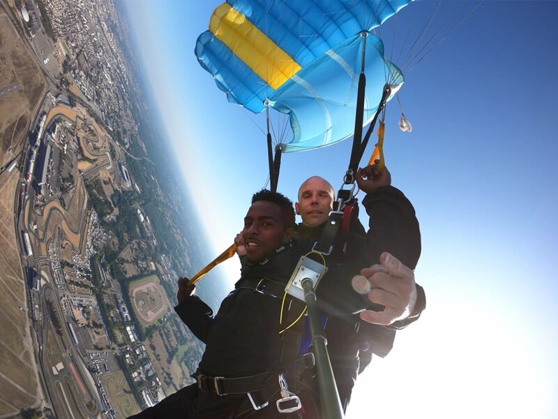 Parachutisme tandem avec un parachute bleu et jaune déployé, vue plongeante sur un circuit automobile et des paysages urbains en contrebas